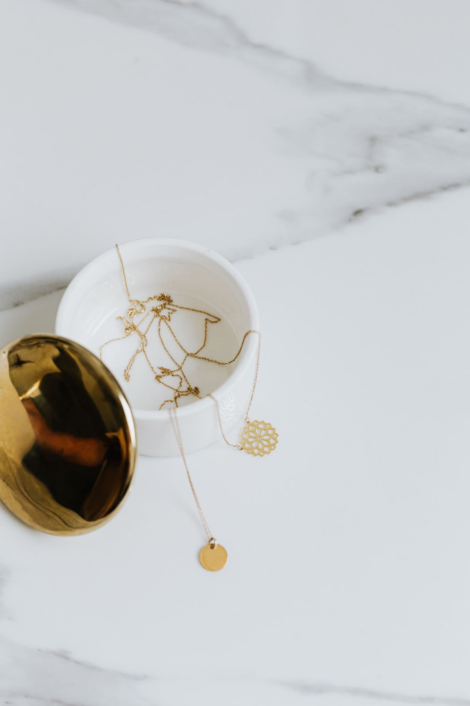 Fine gold chains taken off into a small white dish on a marble surface, the lid set beside it.