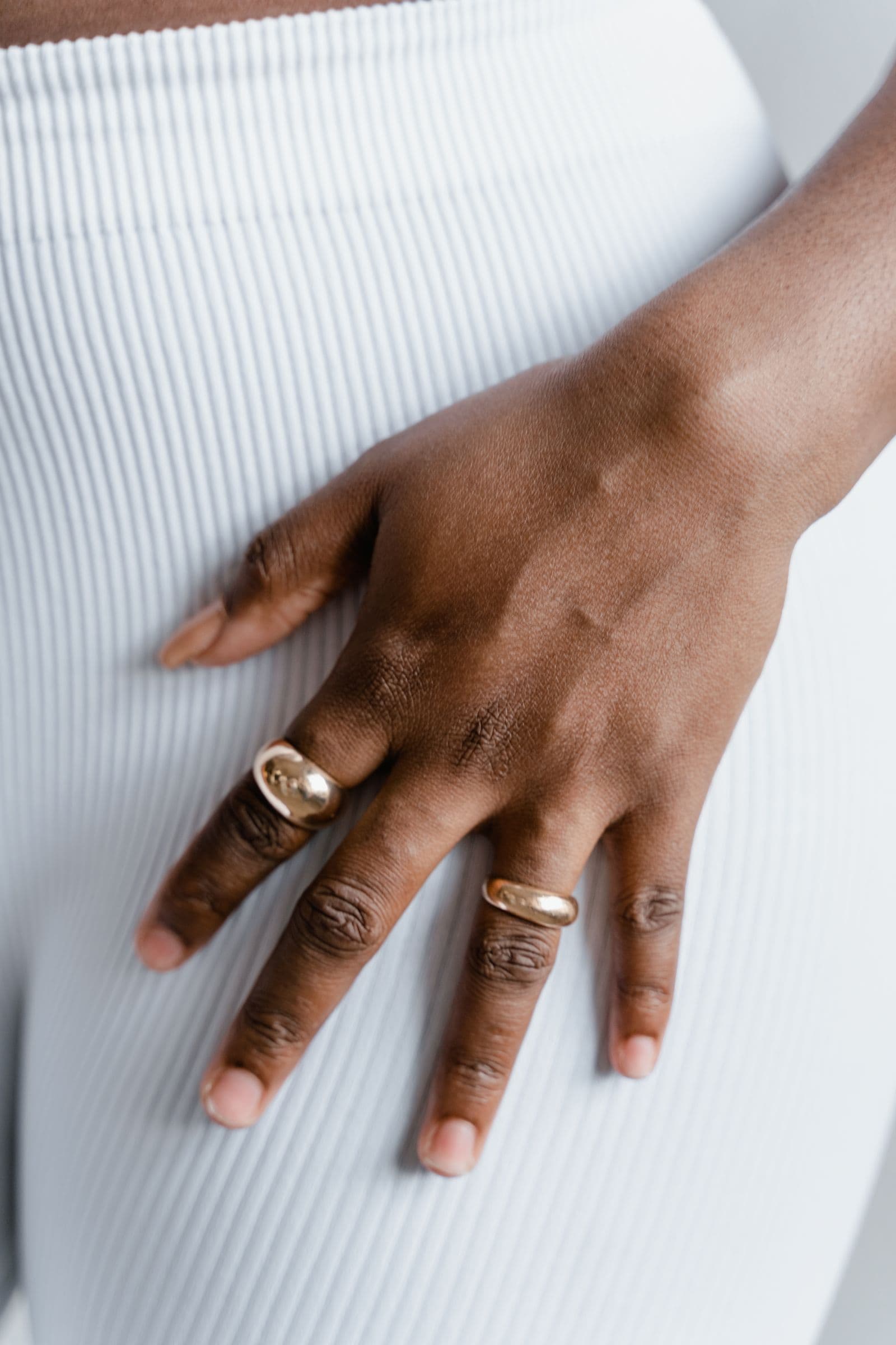 A hand resting against a white ribbed dress, wearing two wide domed gold bands on different fingers.