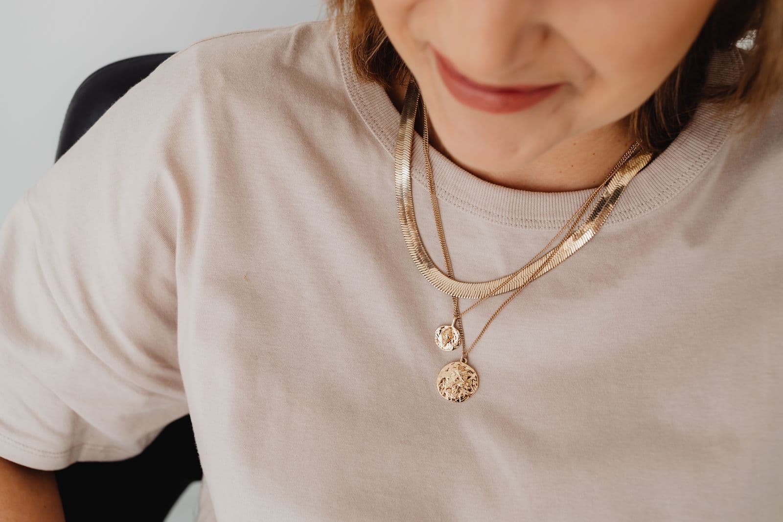 A flat gold chain layered above a finer chain with two coin pendants, worn over a stone-coloured t-shirt.