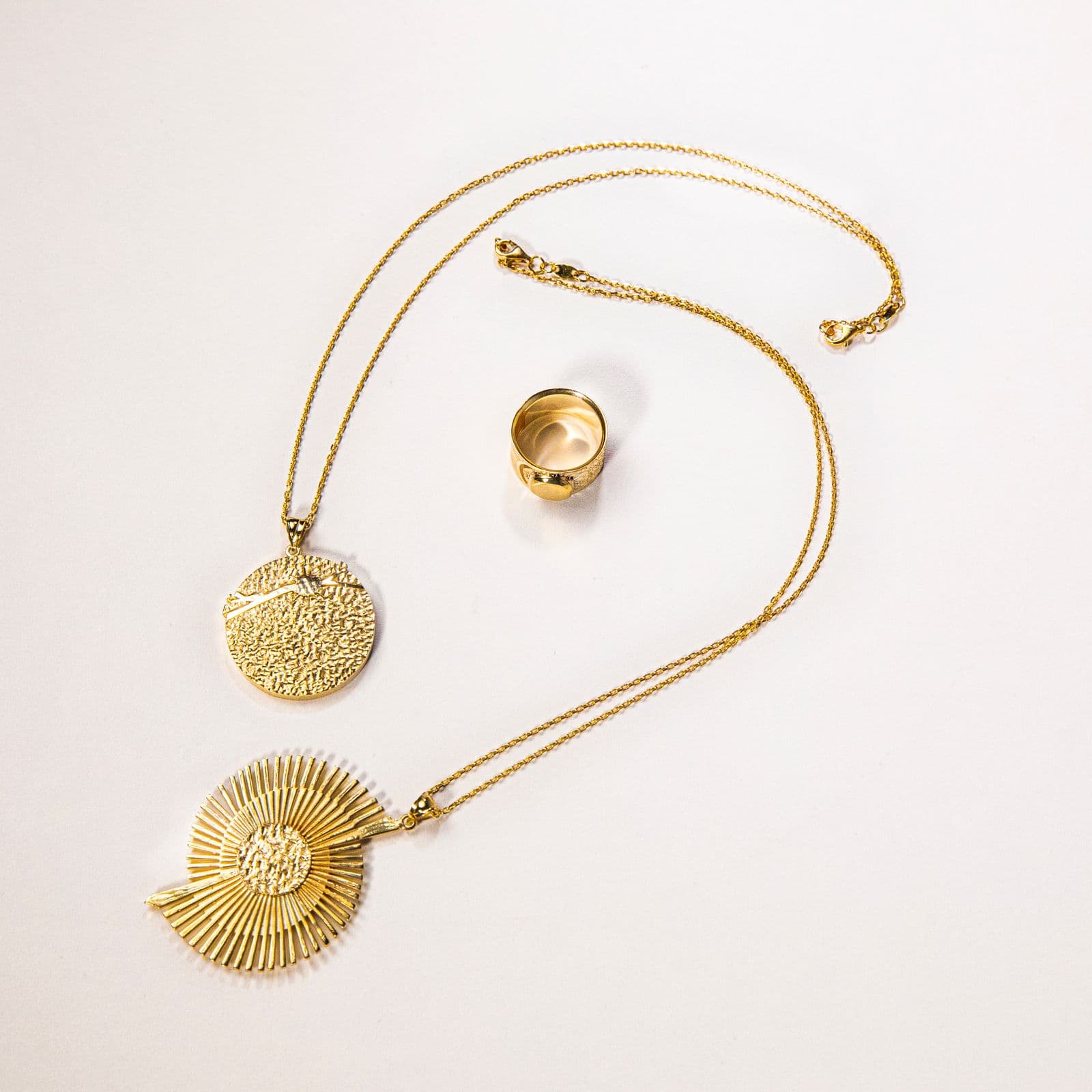 A gold coin pendant necklace, a textured disc pendant and a wide gold band on a pale background.