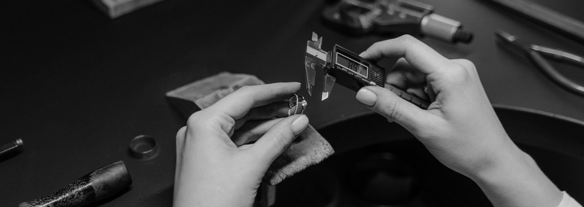A jeweller measuring a finished piece with calipers at the workbench, tools laid out around them.